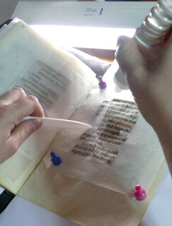 Aerosol wird auf einer handgeschriebenen Seite aufgetragen Eine aufgeschlagene Pergamenthandschrift, Teile davon sind abgedeckt. In einem ausgeschnittenen Bereich wird ein Aerosol appliziert, um die Schrift zu fixieren.